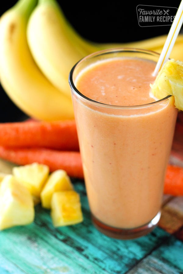 Golden Detox Smoothie in a tall glass with pineapples, bananas, and carrots on the side.