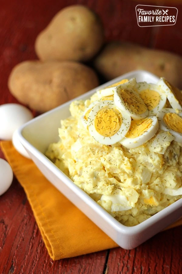 Salade de pommes de terre avec des tranches d'œufs sur le dessus dans un bol blanc et des pommes de terre en arrière-plan