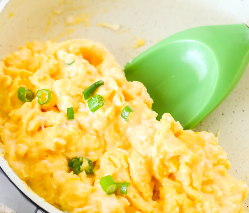 Close up of scrambled eggs in a pan with a green spatula underneath ready to lift the eggs from the pan.