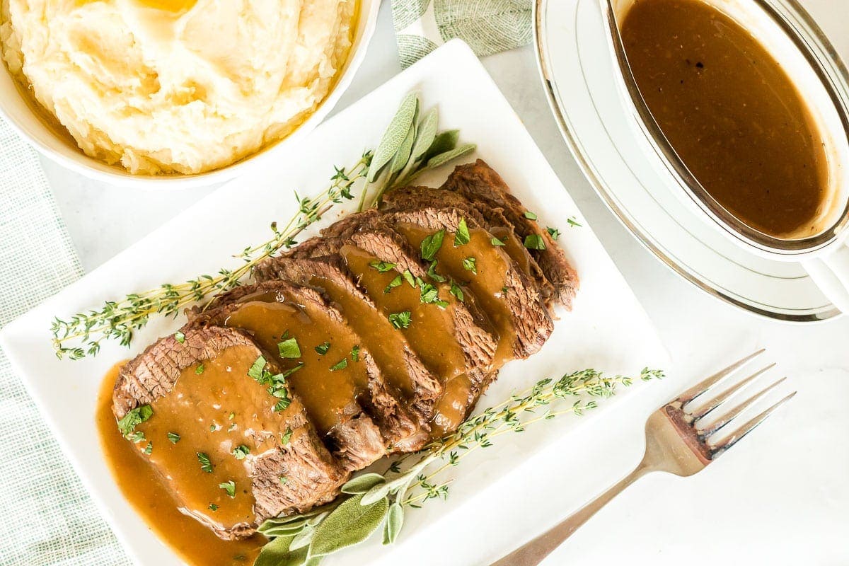 Roast beef on a plate next to a bowl of potatoes and gravy.