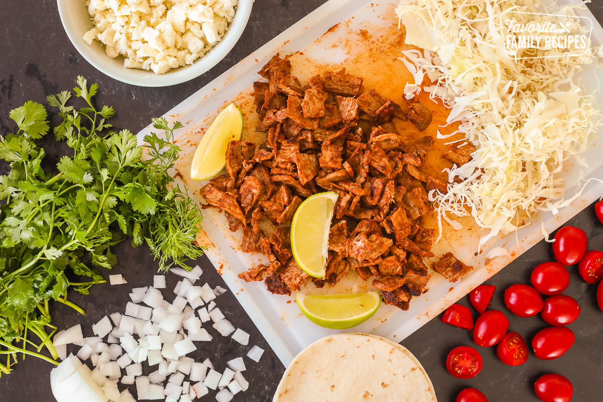 A cutting board with cut grilled steak, limes, shredded cabbage, tomatoes, onion, cheese, and tortillas.