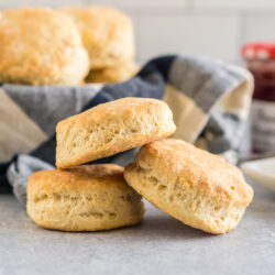 Three buttermilk biscuits stacked on each other with a stick of butter and a jar of jam in the background