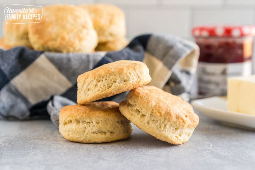 Three buttermilk biscuits stacked on each other with a stick of butter and a jar of jam in the background