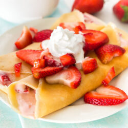 Two crepes on a white plate with strawberry filling and topped with whipped cream and strawberries