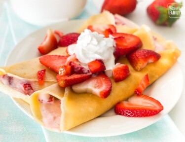 Two crepes on a white plate with strawberry filling and topped with whipped cream and strawberries