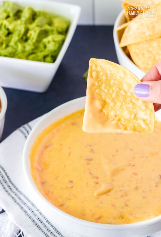 Hand holding a dipped tortilla chip over a bowl of Nacho Cheese Dip. Chips and guacamole on the side.