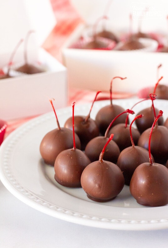 Close up plate of Chocolate Covered Cherries with gift boxes of cherries behind.