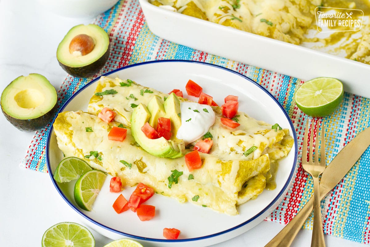 Plate of two Spinach Enchiladas topped with tomatoes, avocados, limes next to baking dish of enchiladas.