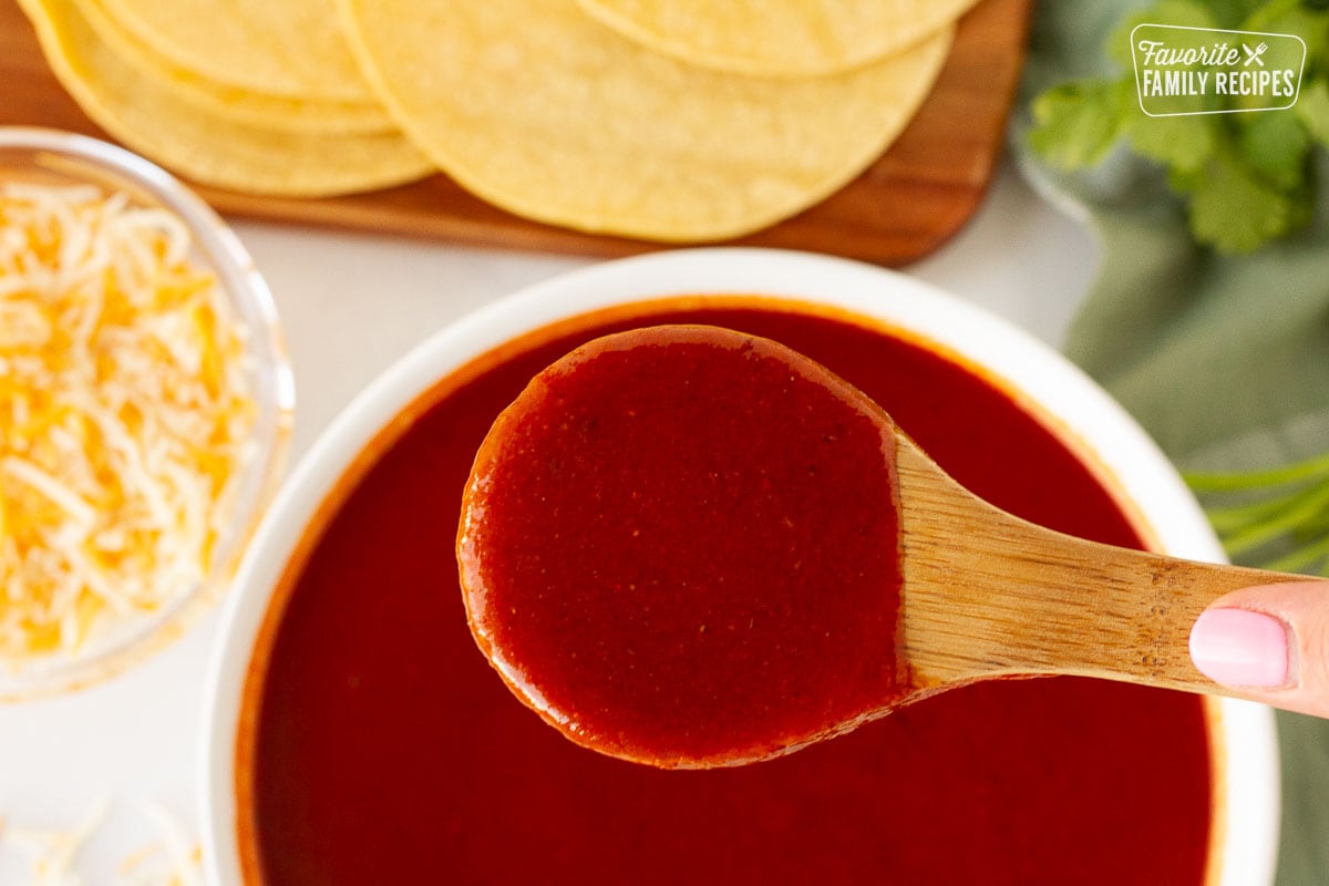 Hand holding up a wooden spoon full of Homemade Enchilada Sauce over a bowl of sauce. Tortillas, cilantro and shredded cheese on the side.