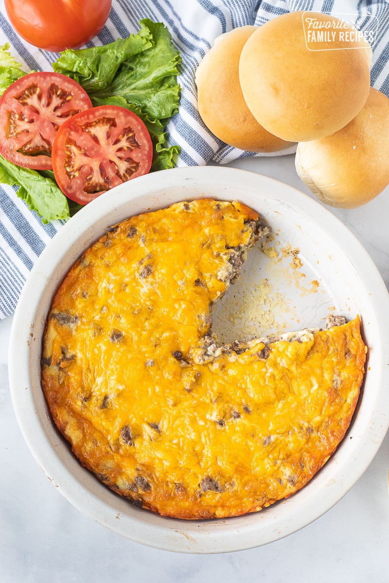 Pie plate missing a slice of Cheeseburger Pie. Lettuce, tomatoes and rolls on the side.