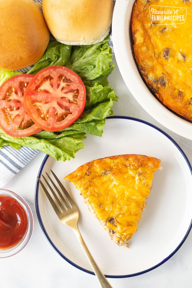 Slice of Cheeseburger Pie on a plate with ketchup, sliced tomatoes, lettuce and buns on the side.