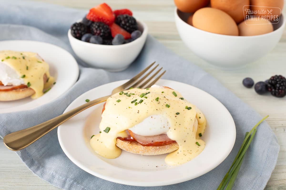 two plates of eggs benedict with a bowl of berries and a bowl of eggs in the background