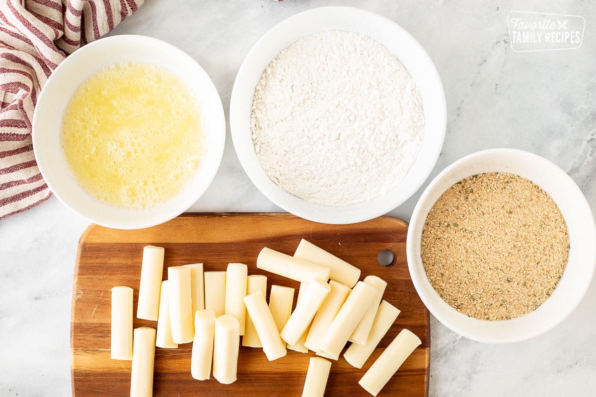 Cutting board with halved string cheese. Bowl of egg whites, bowl of flour mix and bowl of bread crumbs.