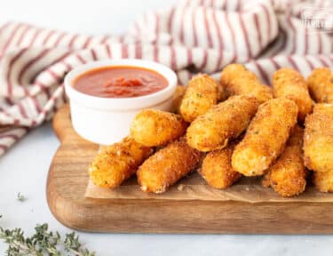 Homemade Mozzarella Sticks stacked on a board with a cup of marinara sauce.