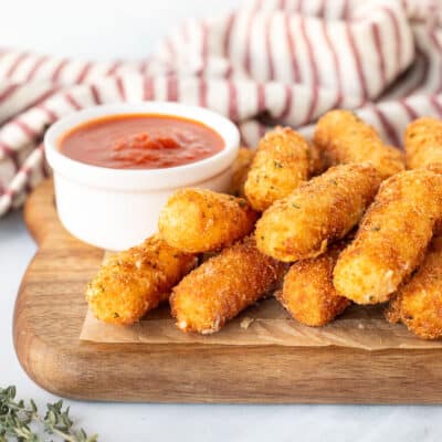 Homemade Mozzarella Sticks stacked on a board with a cup of marinara sauce.