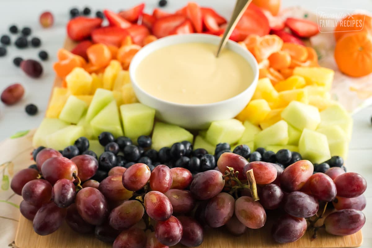 Cream cheese fruit dip with a spoon on a platter with strawberries, orange slices, pineapple, honeydew, blueberries, and grapes