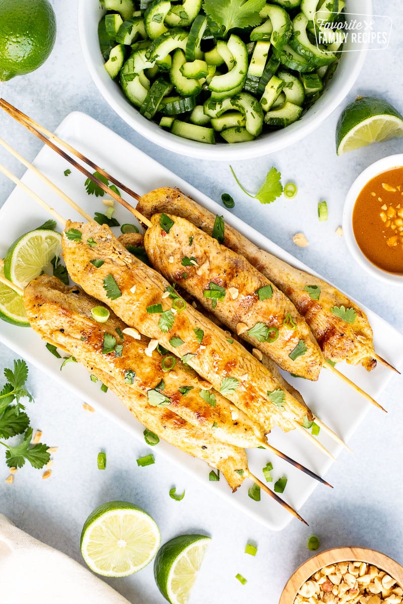 Chicken Satay on a glass rectangular dish on wooden skewers. Garnished with lime wedges, cilantro, green onion and peanuts.