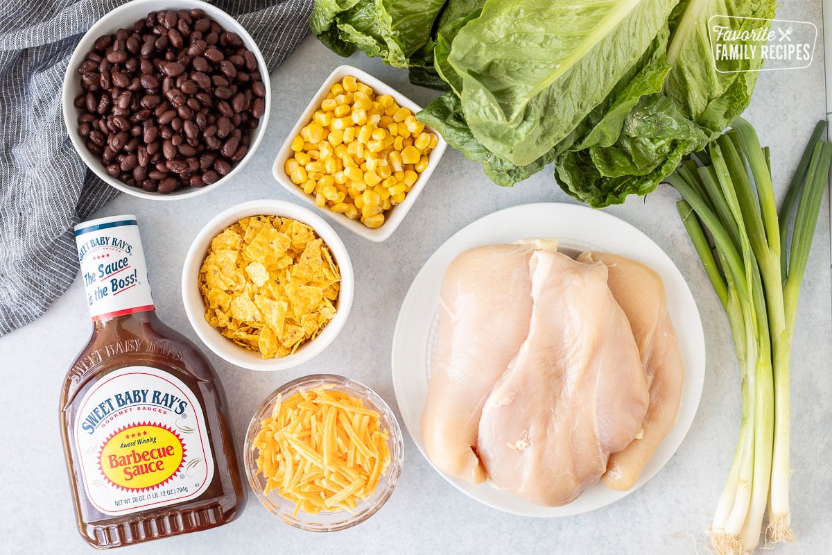 Ingredients to make Barbecue Chicken Salad including lettuce, chicken breasts, green onion, corn, black beans, cheddar cheese, crushed tortilla chips and barbecue sauce.