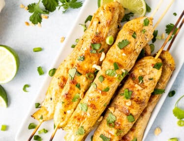 Plate with Chicken Satay garnished with cilantro, green onions and peanuts. Sliced limes on the side.