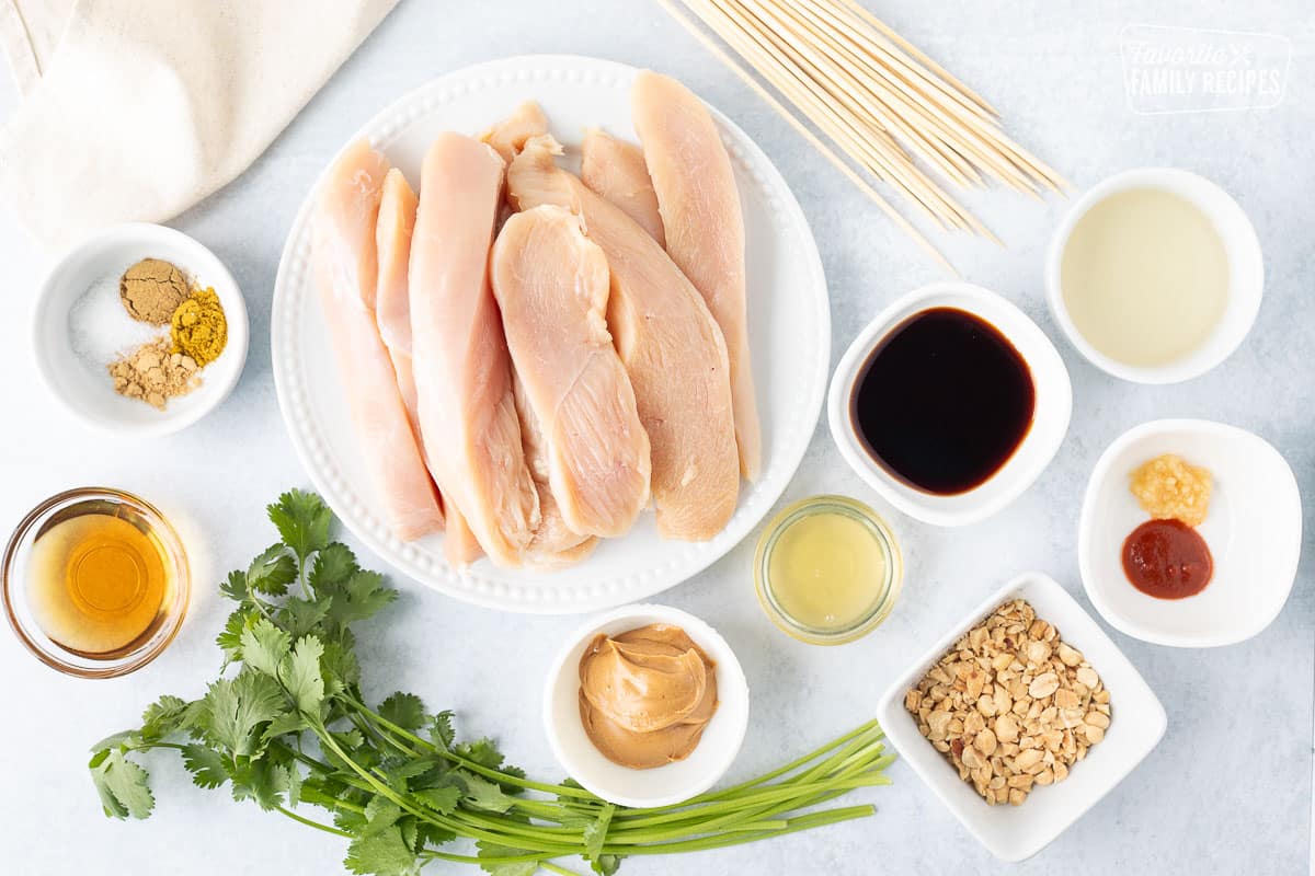 Ingredients to make Chicken Satay including sliced chicken breasts, wooden skewers, cilantro, soy sauce, rice vinegar, garlic, sriracha, peanuts, peanut butter, honey, spices and peanut oil.