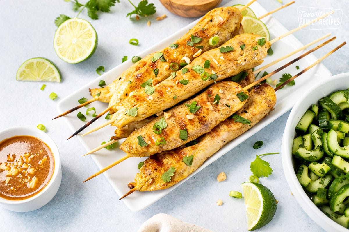 Chicken Satay on a glass plate. Peanut sauce on the side. Bowl of Thai cucumber salad on the side.