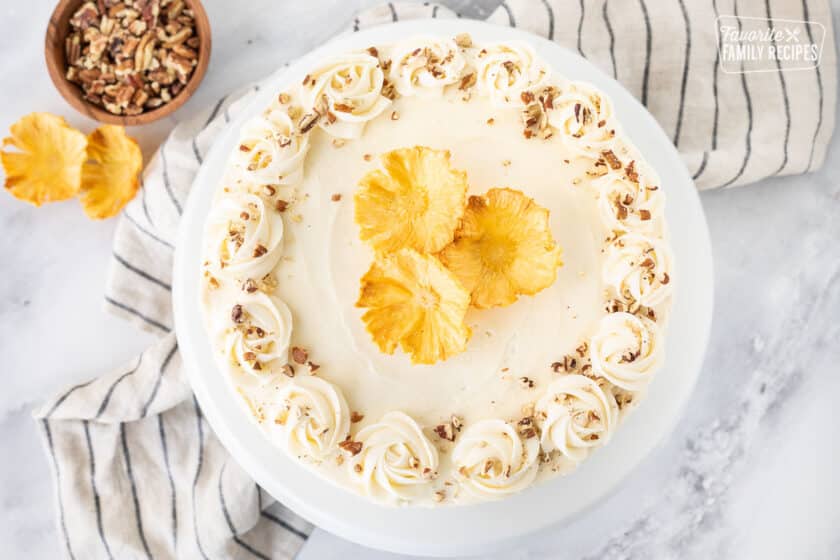 Whole Hummingbird Cake from the top view on a cake stand. Topped with cream cheese frosting, toasted pecans and dried pineapple flowers.