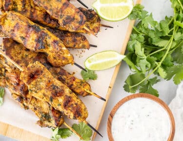 Grilled Thai chicken skewers on a wooden board with cucumber yogurt dip.