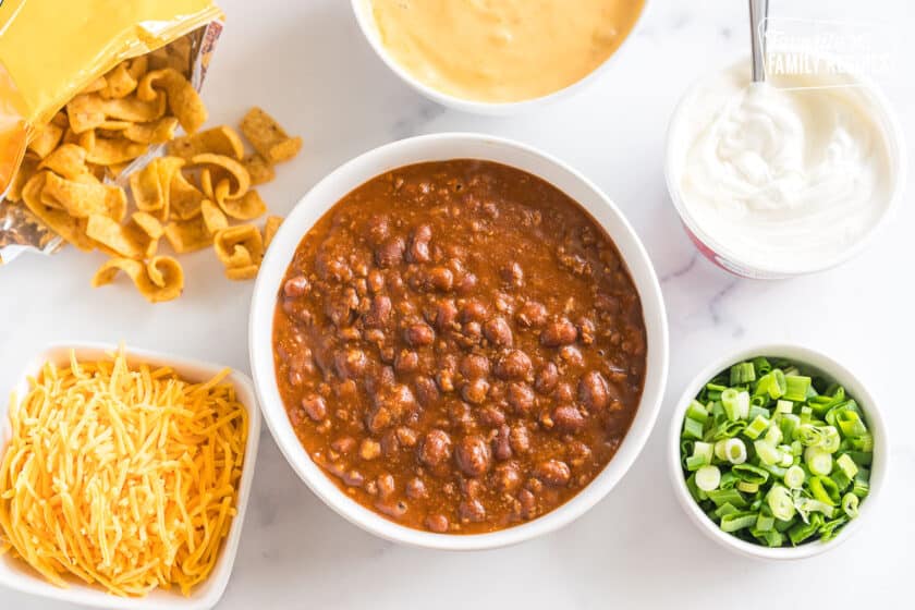 Bowls of chili, queso, sour cream, cheese, green onions, and Fritos on the counter