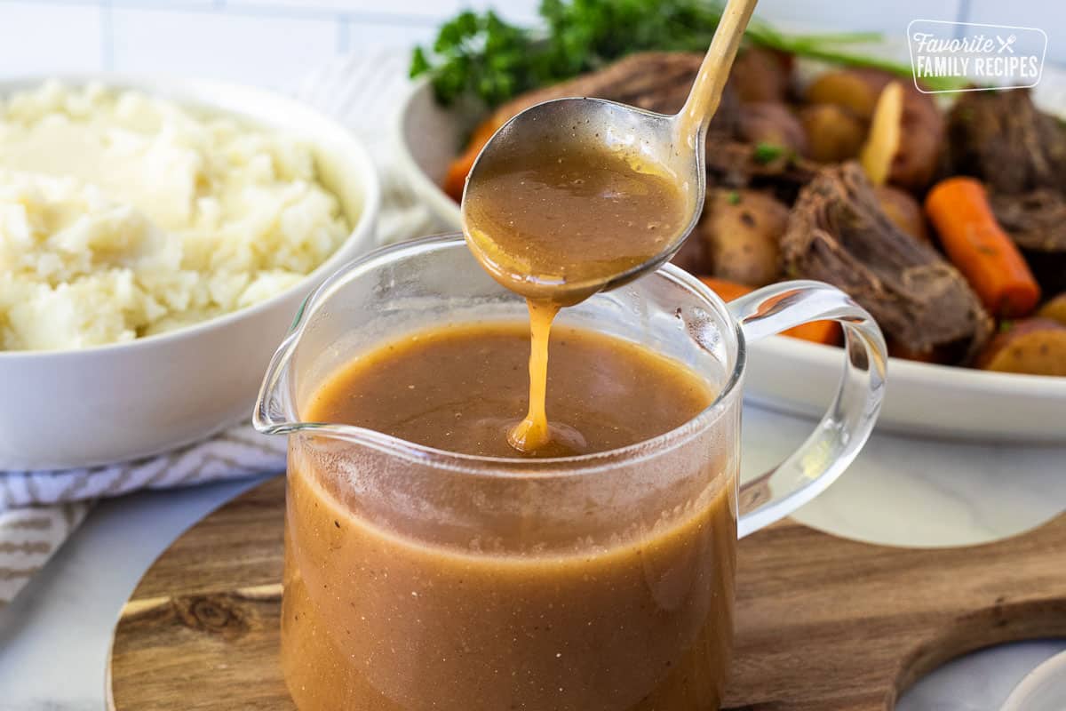 Beef Gravy poured from a ladle into glass jar. Mashed potatoes and beef roast in the background.