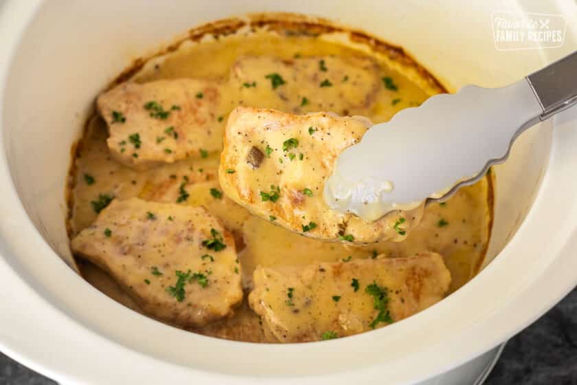 Tongs holding a Pork Chop above a crock pot with cooked crock pot pork chops and gravy.