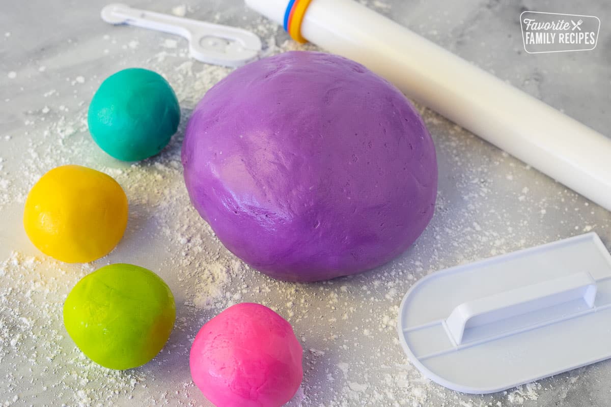 Large ball of purple fondant with small balls of teal, yellow, lime green and hot pink fondant. Roller, cutting a smoother on the side.
