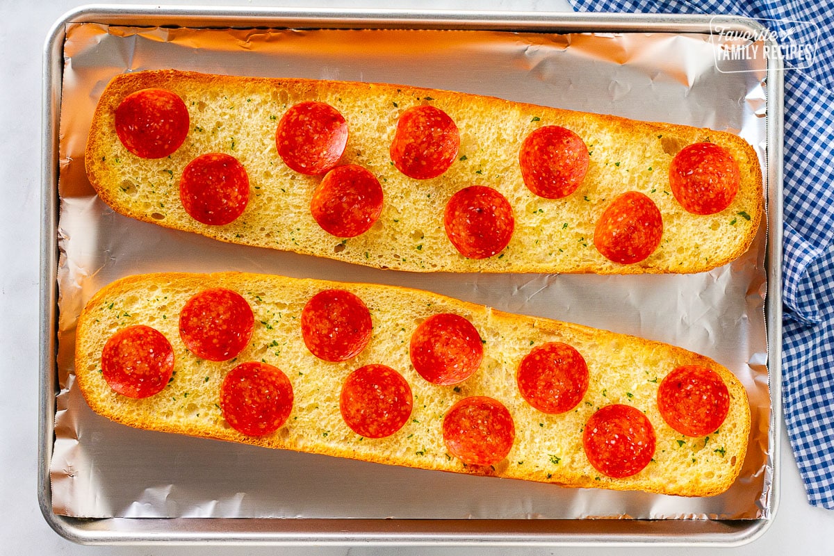 Two halves of French bread on a sheet toasted with butter and topped with pepperoni.
