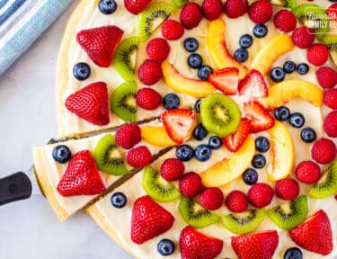 Whole fruit pizza with a slice cut and held with spatula.