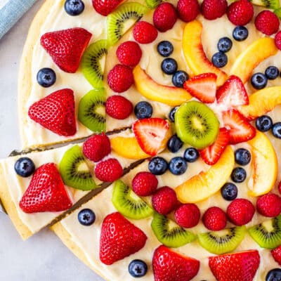 Whole fruit pizza with a slice cut and held with spatula.