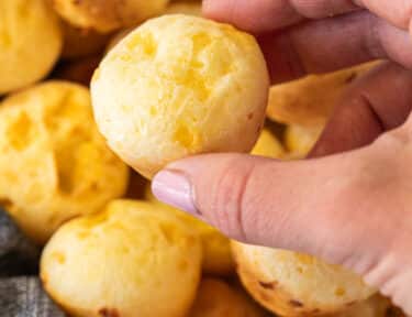 Hand holding a Brazilian Cheese Bread roll above a basket of golden rolls