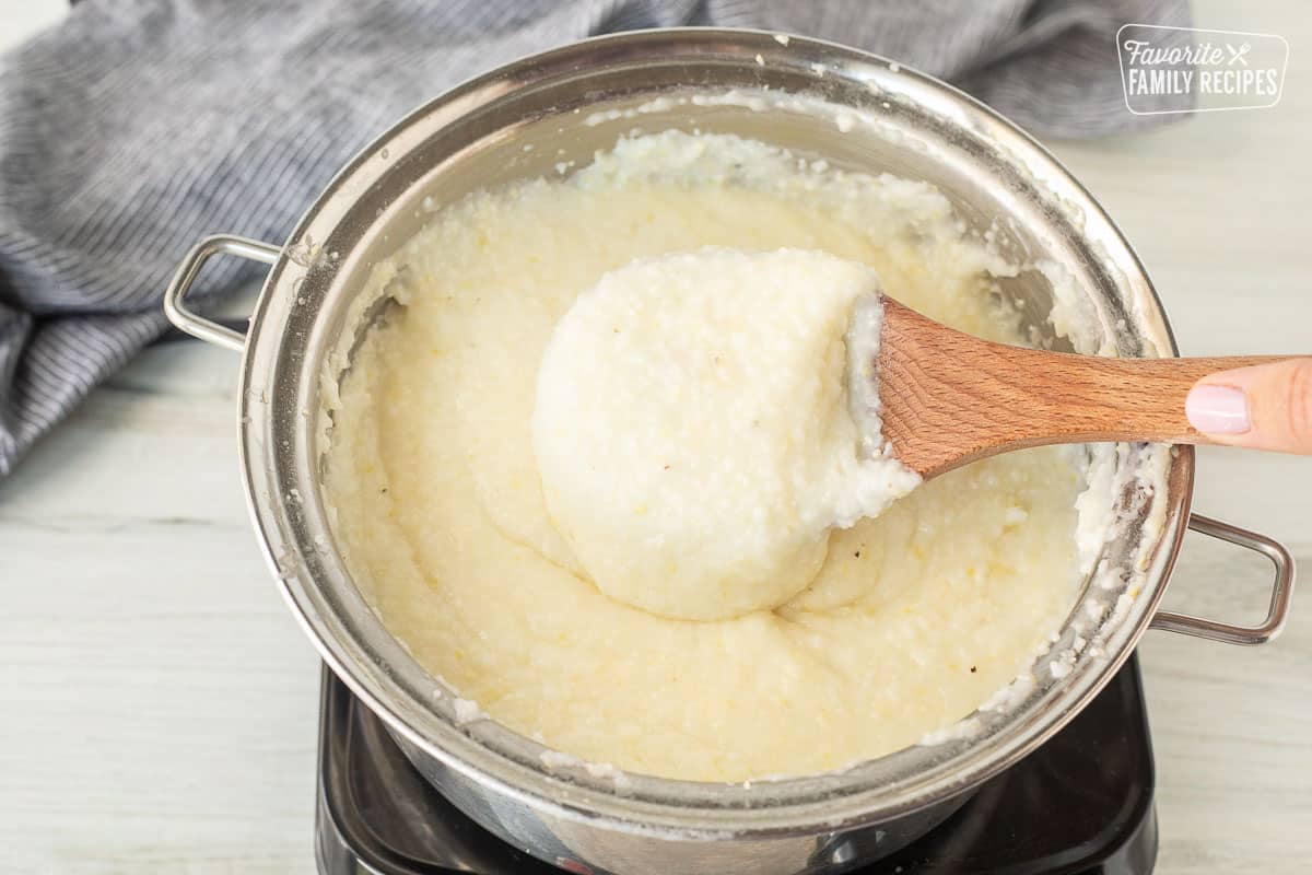 Wooden spoon holding up cooked grits in a saucepan.