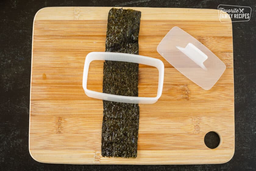 A strip of Nori roasted seaweed under a Spam Musubi mold on a cutting board.