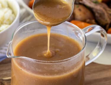 Pouring Beef Gravy into a glass container of gravy. Mashed potatoes and vegetables in the background.