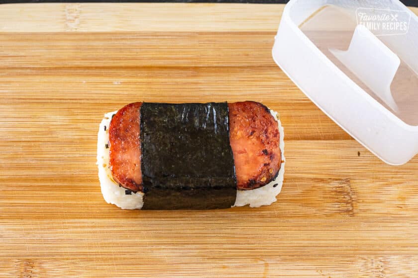 Spam Musubi Hawaiian snack next to a musubi mold on a cutting board to demonstrate the shaping process.