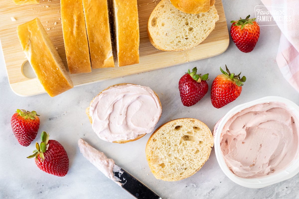 Spreading strawberry cream cheese on a slice of French bread with a knife.