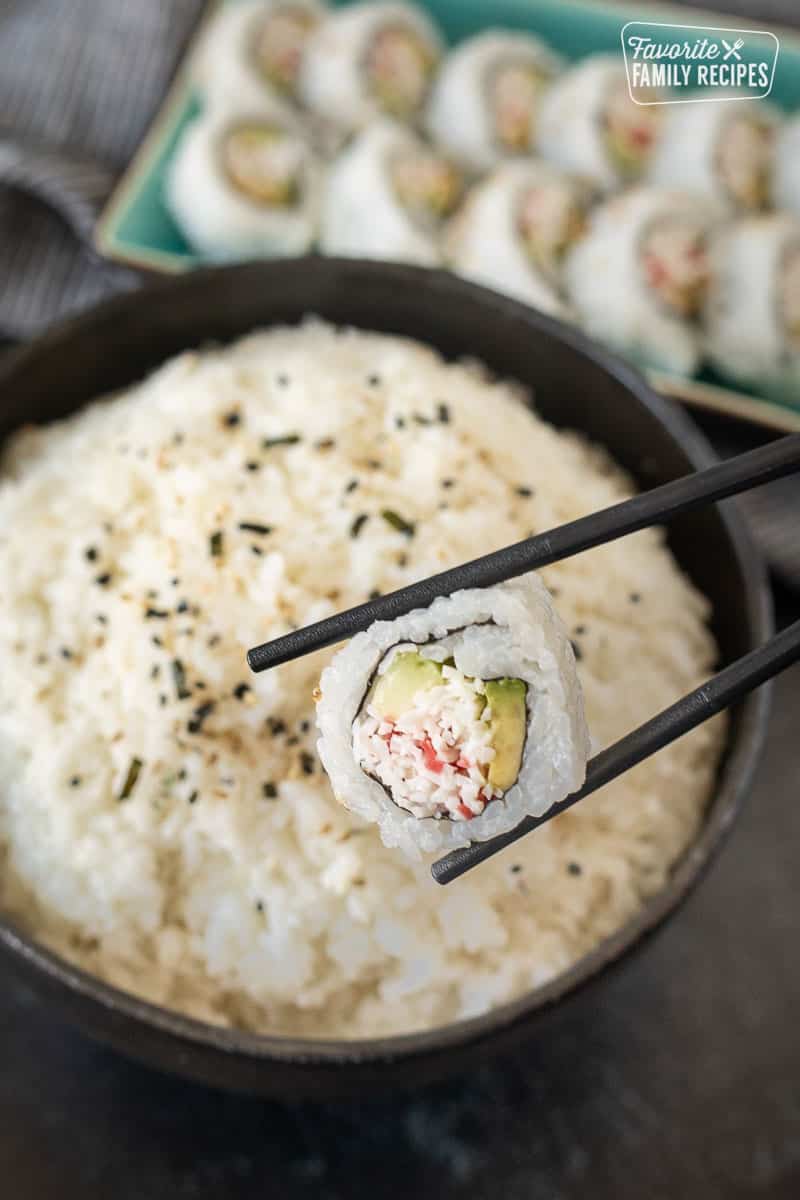Chop sticks holing a sushi roll over a bowl of sushi rice.