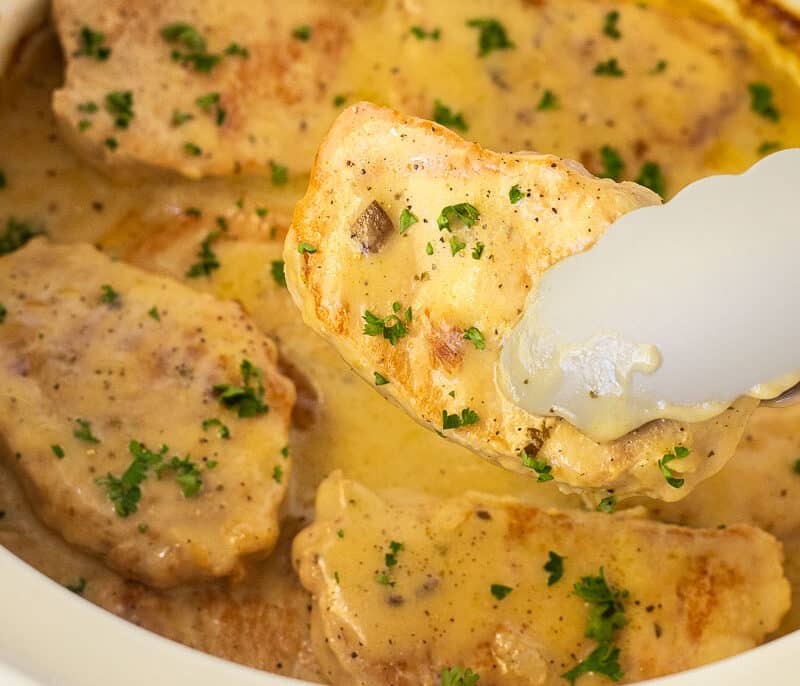 Tongs holding a Pork Chop above a crock pot filled with cooked, tender crock pot pork chops.