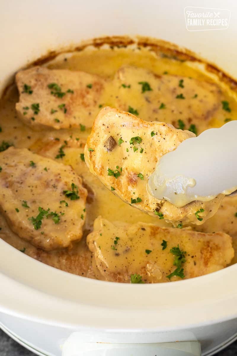 Tongs holding a Pork Chop above a crock pot filled with cooked, tender crock pot pork chops.