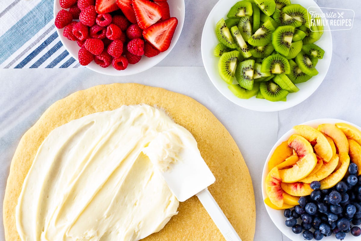 Cookie pizza with a spatula spreading cream cheese frosting and three bowls of fresh fruit on the side.