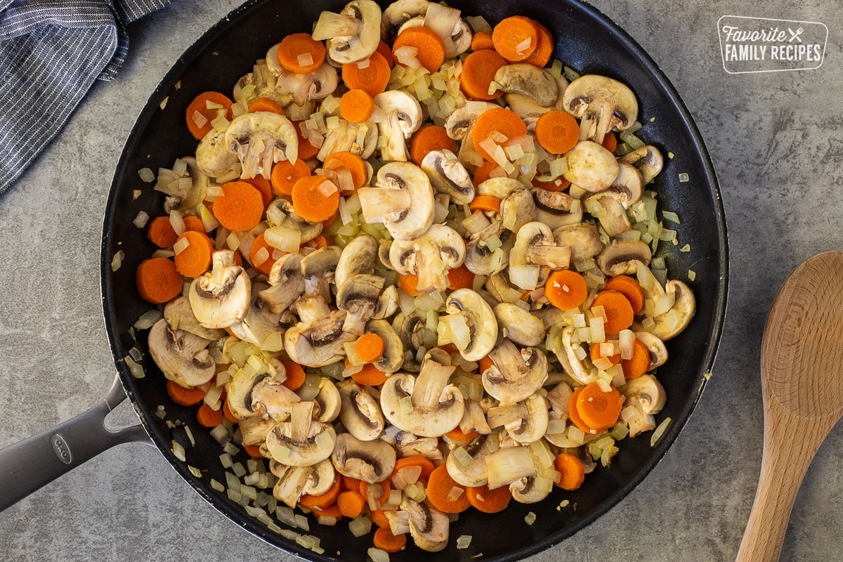 Skillet with sautéed mushrooms, carrots and onion. Wooden spoon on the side.