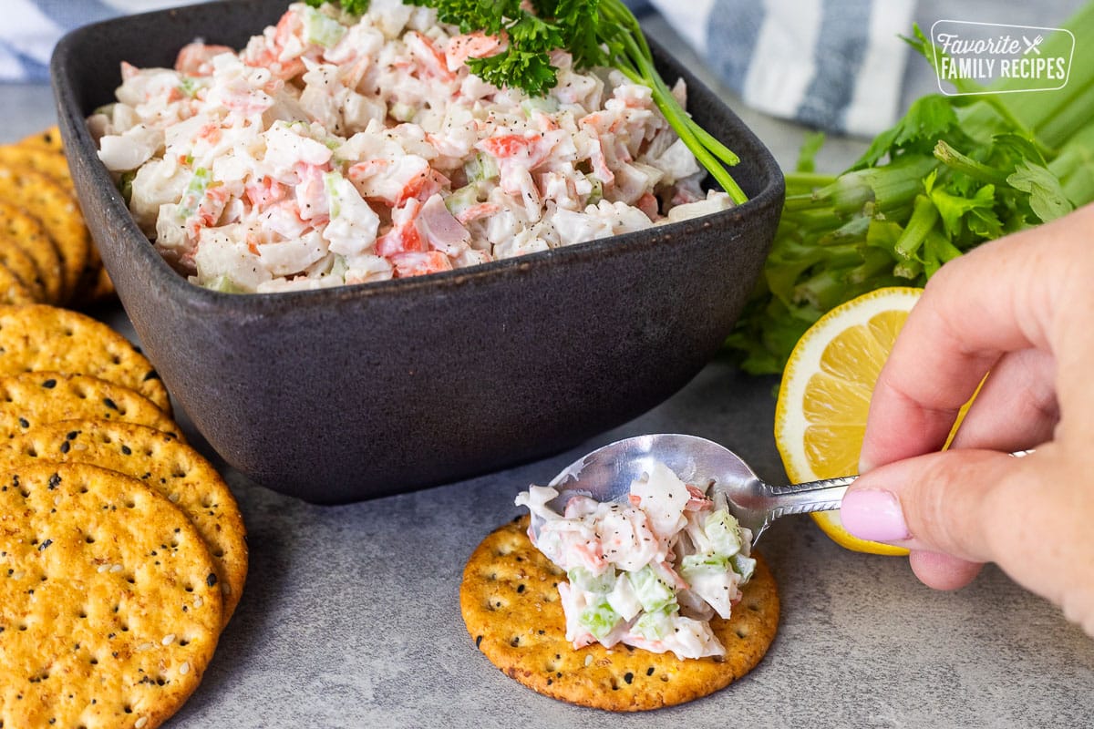Hand placing Cold Crab Dip on a cracker. Bowl of cold crab dip and extra crackers in the background.
