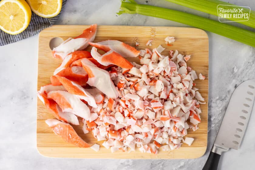 Cutting board with cut up imitation crab. Knife, celery and lemons on the side.