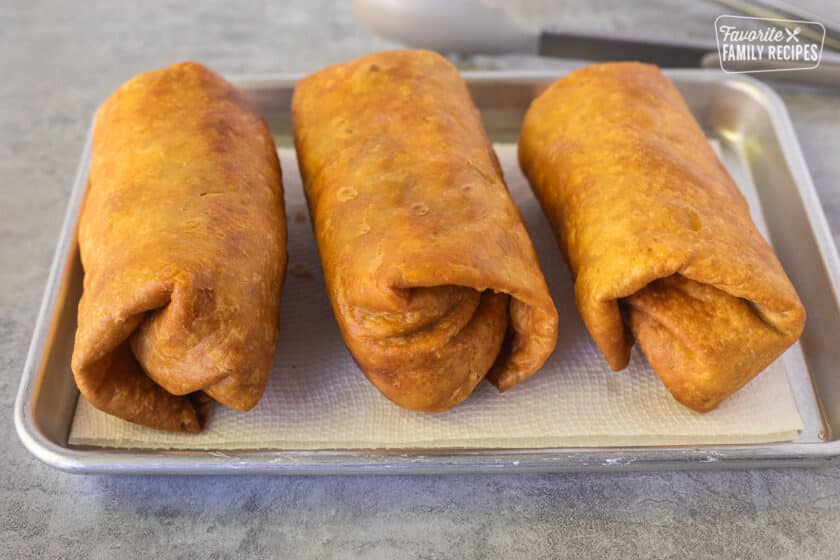 Three fried Chicken Chimichangas on a paper towel on a baking dish.