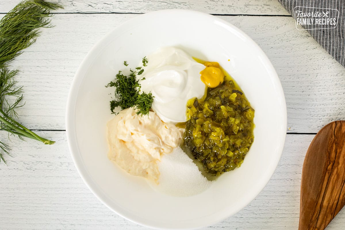 Glass bowl with relish, mayo, sour cream, dill, sugar and mustard. Wooden spoon on the side.