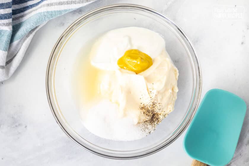 Glass bowl of ingredients for macaroni salad dressing including mayo, sour cream salt, sugar, pepper, mustard and apple cider vinegar. Spatula on the side.